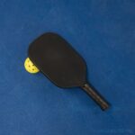 A black pickleball paddle resting on a bright yellow ball on a blue surface