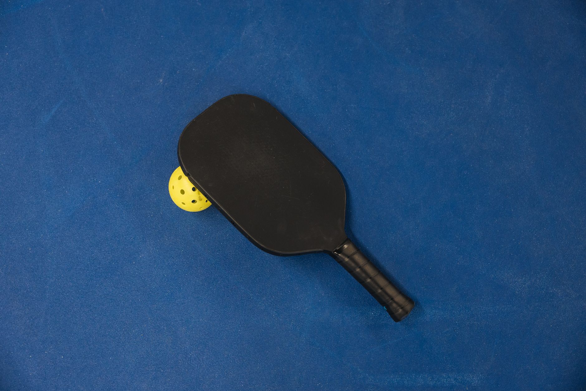 A black pickleball paddle resting on a bright yellow ball on a blue surface