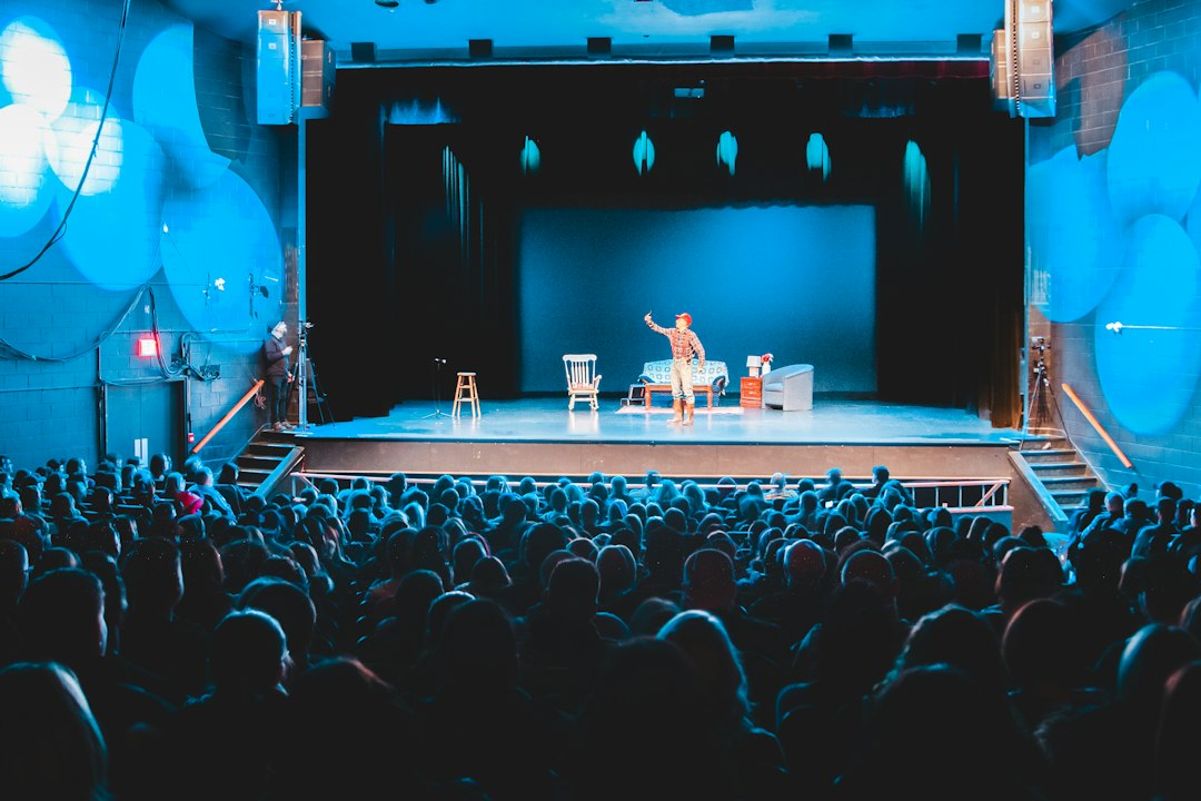 A performer on stage in front of a large audience, with various stage props visible.
